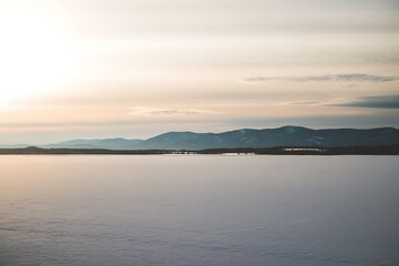 A tranquil winter scene snowcovered lake reflecting a calm sunset, with distant mountains silhouetted against the soft light. Peaceful and serene.