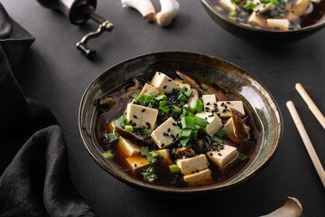 Fresh traditional miso soup with tofu, shiitake mushrooms, green onion, wakame and spices on black stone background top view. Traditional Japanese cuisine.