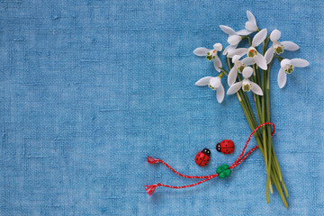 Bouquet of snowdrop flowers and red and white cord, ladybugs on a blue fabric background. Postcard for the holiday of March 1, Martisor, Baba Marta