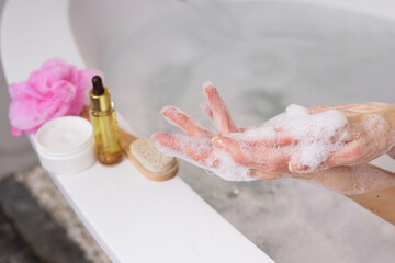 Relaxing spa moment. Woman washing hands with foamy soap near a bathtub. Skincare essentials like cream, oil, brush, and loofah on the side. Self-care, hygiene, and wellness concept