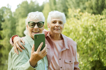 Two Caucasian pretty senior women smiling to smartphone camera while taking selfie photos near lake in the park.