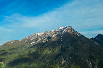 Iconic transmitter tower atop snow-capped Dobratsch mountain stands tall against clear blue sky. Jagged rocks and patches of evergreen forest cling to slopes, revealing rugged terrain of Gailtal Alps