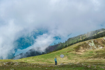 Hiker woman walks on Mirnock grassy slopes gazing towards cloud-veiled Lake Millstatt in Carinthia, Austria. Alpine scenery blends with distant, misty waters. Serene alpine landscape and green pasture