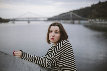Fototapeta premium Pensive young woman near the river on the background of the city river.