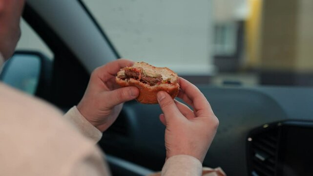 Man takes bite of tasty burger to quell hunger sitting in car on cloudy day. Man consuming burger in car and enjoying view from window. Man indulging in unhealthy burger in car in move on road of city