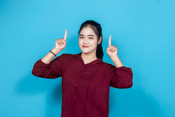 Smiling young woman enthusiastically pointing with fingers against a vivid blue backdrop, dressed...