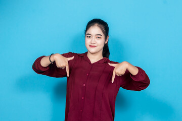 Smiling young woman enthusiastically pointing with fingers against a vivid blue backdrop, dressed...