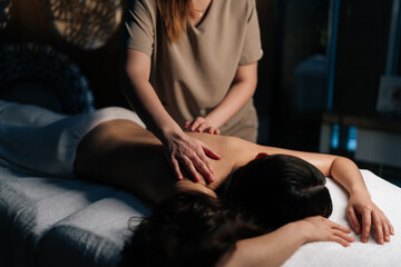 Obraz premium Closeup of unrecognizable masseuse applying massage oil back of young woman lying on massage table, providing relaxing and therapeutic body treatment in spa salon with dark interior.