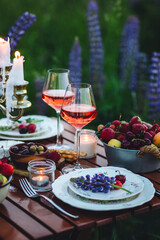 Romantic table decor for a loving couple on blooming meadow with purple lupines. Two glasses of wine, flowers in a vase, silverware, fruits, wooden furniture. Sunset, golden hour, atmospheric light