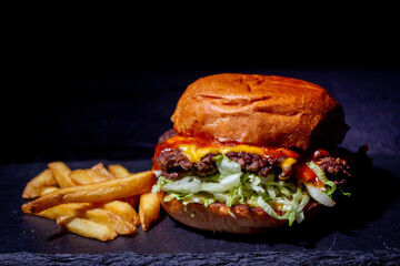 Very delicious burger , with juicy meat, salad, a slice of  cheese and ketchup, with a golden crust and golden fries on the side on the black stone table