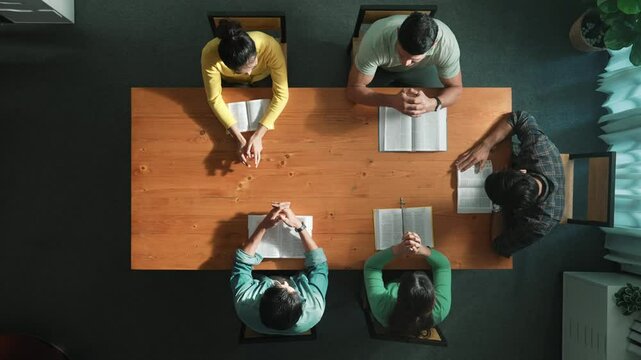 Top down aerial view of smart team hold hands on the holy bible and praying god while sitting at meeting table. Group of people hand folded in prayer with faith, spirituality and religion. Symposium.