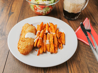 Popular fish and chips meal on a white plate and brown wooden table. Chips are made from sweet potatoes and cut in long chunks. There is splash of sous or cream on top of fine food.