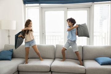 Two playful children holding pillows, engaged in active pillow fight while standing on large sofa in modern light living room. Siblings having fun together on weekend leisure at home, games, childhood