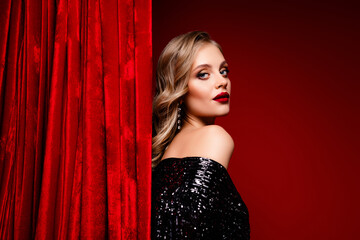Tempting blonde woman in stylish black sequin dress against red velvet curtain background