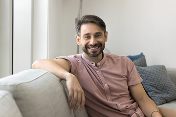 Fototapeta premium Portrait of smiling middle-aged Portuguese man dressed in casual shirt relaxing on comfortable couch in modern living room, looking, posing for photo, feel satisfied, spending carefree pastime at home