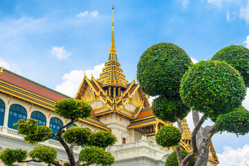 Grand Palace in Bangkok, Thailand, featuring traditional Thai architecture with golden roof, ornate details, and manicured green trees against bright blue sky. Travel and touristic landmark in Asia