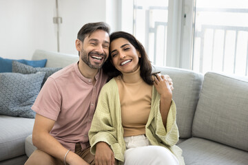 Obraz premium Young beautiful multiethnic couple sits comfortably on sofa, laughing, enjoying each other company, spending carefree leisure and tender moments together at modern home. Strong marriage, bond, love