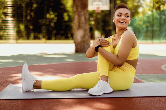 Laughing black woman stretching legs outside. African American female athlete in wireless earbuds training abductor hip flexor muscle in buttocks to relieve sports pain practicing seated spinal twist