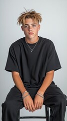 A young man with a shaved head and a black shirt sits on a black chair. He is wearing a silver necklace and a bracelet. Concept of confidence and self-assurance