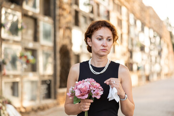 Fototapeta premium Elegant grieving woman in forties, wearing formal black dress and pearls, standing with bouquet in hands near stone columbarium wall in cemetery, gazing sorrowfully and contemplating loss