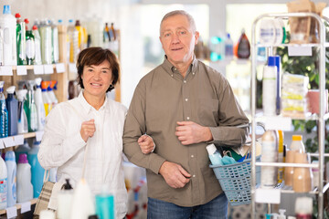Obraz premium Elderly couple with a basket full of purchases chooses household chemicals in the supermarket. Shoppers look at the shelves of home cleaning products