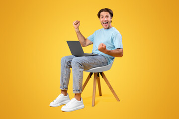 Online Win. Excited Happy Guy Sitting On Chair With Laptop Computer, Emotional Young Man Celebrating Success Shaking Clenched Fists Over Yellow Studio Background, Full Length Shot With Free Space