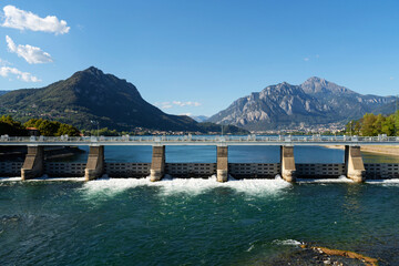 Large dam structure spans across lake with water flowing through the gates at lake Como, near Lecco, Italy. Towering mountains under clear blue sky with small boat visible on the lake