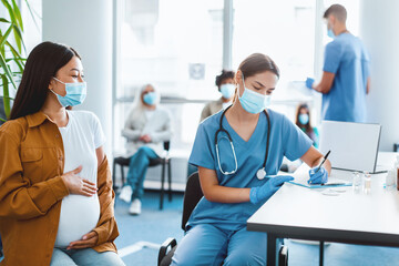 Obraz premium Mass Vaccination. Woman doctor in medical mask using clipboard, writing while talking with pregnant patient prepared to get vaccinated in hospital, getting immunization jab shot against corona virus