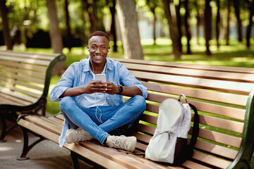 Obraz premium Cool black guy in earphones listening to music online from his smartphone on bench at summer park, empty space