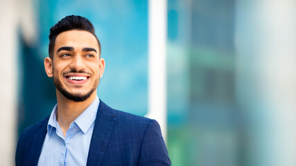 Fototapeta premium Closeup portrait of handsome young bearded arab man in suit entrepreneur or manager posing by modern business center, smiling and looking at copy space. Business opportunities concept