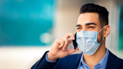 Closeup portrait of young arab businessman in protective medical face mask talking on cellphone next to business center, copy space. Muslim entrepreneur or CEO working during COVID-19 pandemic