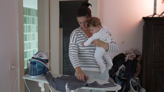 Mother ironing clothes while holding baby, multitasking in daily routine, balancing household chores with parenting, authentic family moment, nurturing care, candid domestic life indoors