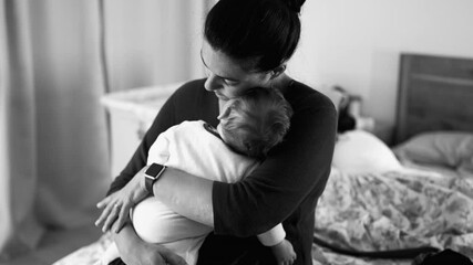 Mother holding baby tenderly in bedroom, providing comfort and emotional support through close embrace, warmth and bonding between parent and child in black and white
