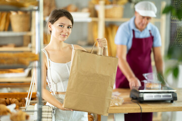 European positive young girl holds out paper bag with pastries in hand in small bakery store