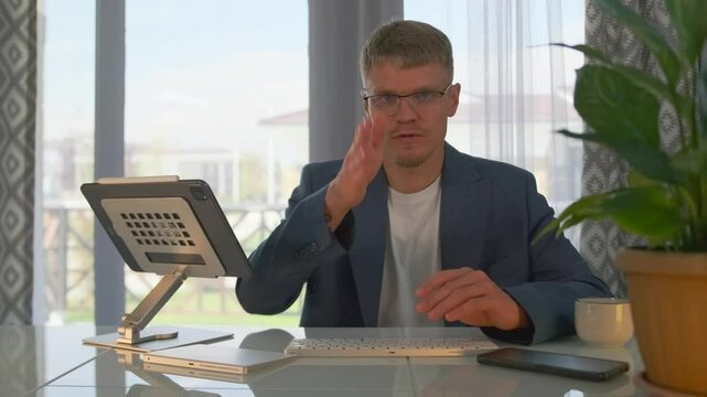 The tutor conducts a webinar, explaining concepts and engaging with participants using a tablet. He gestures while speaking, responding to questions, in a well lit room with a panoramic view.