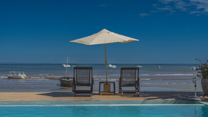 Two sun loungers in the shade of a sun umbrella by the swimming pool.  The ocean is ahead. Low tide. An old fishing boat in shallow water. The motorboats are far away. The blue sky. Madagascar.Nosy Be