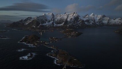 A breathtaking aerial view of the Lofoten Islands in Norway, featuring a stunning coastal landscape with snow-capped mountains at dusk. The scattered lights reflect on the water,