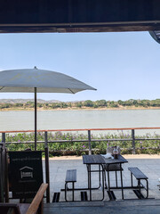 serene view of river from restaurant, featuring table and chairs under umbrella, surrounded by greenery and mountains in distance. peaceful atmosphere invites relaxation and enjoyment