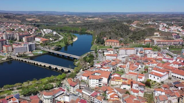Scenic panorama of Mirandela cityscape on banks of Tua overlooking residential districts, two bridges across river and gentle mountain slopes covered with forests in background on spring day, Portugal