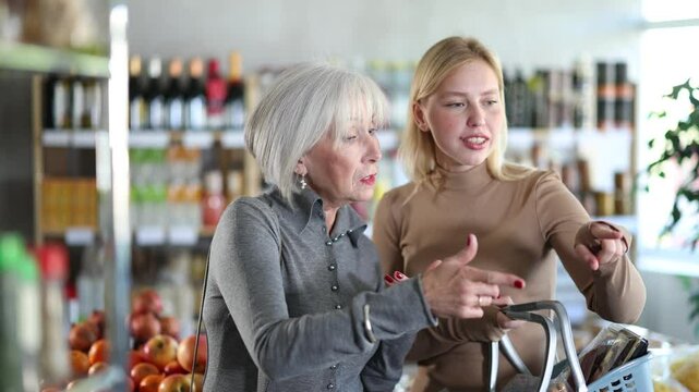 In shop minimarket, family young woman and grandma stares at windows. Client holds shop basket of groceries and foodstuff in hands, reaches for shelf, trying to see products in refrigerator window