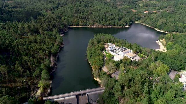 Drone shot of a dam and the respective river from both sides. There is also a building by riverside, and many kilometers of forest, that extends into every direction.