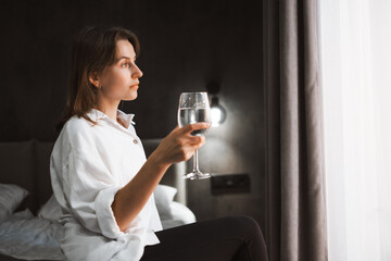 Woman sits on the edge of cozy comfortable hotel bed, looking out the window dreaming with glass of water. Person reflecting on travel experiences and plans, surrounded by her luggage.