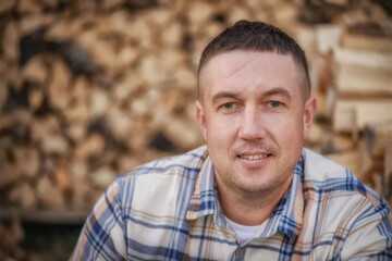 Portrait of a handsome young man in a checkered shirt and beige breeches. The man is sitting against the background of wooden logs. A man in the village. The man chopped logs. The man is sitting on a 