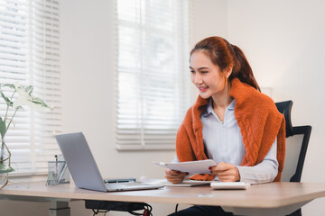 Woman working at desk with laptop, holding documents and smiling in bright office.