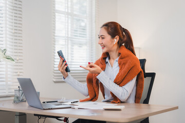 Obraz premium Woman in home office smiling while using smartphone and wearing wireless earbuds.