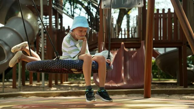 Young boy wearing bucket hat sitting pensively on playground swing, gazing into distance with contemplative expression amid colorful recreational space during sunny summer day