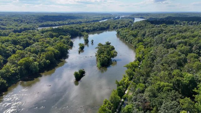 A cinematic drone shot pans up, showcasing the Potomac River. The water's edges curve gracefully, with trees guiding the flow, creating a natural, serene landscape under a bright sky.