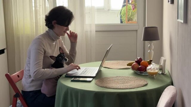 
Almaty, Kazakhstan - 02.02.2025 : A woman is sitting at her laptop, with her dog, in the kitchen. Work from home.