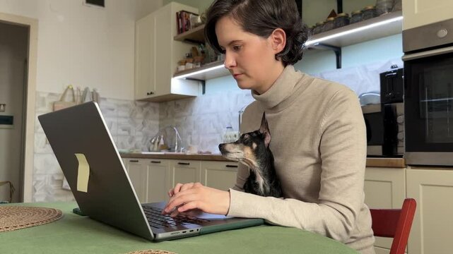 
Almaty, Kazakhstan - 02.02.2025 : A woman is sitting at her laptop, with her dog, in the kitchen. Work from home.