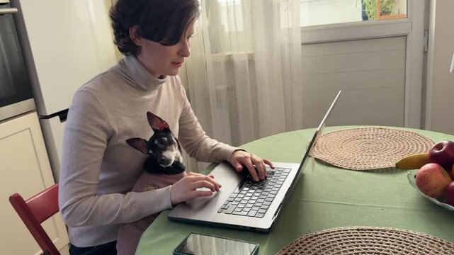 
Almaty, Kazakhstan - 02.02.2025 : A woman is sitting at her laptop, with her dog, in the kitchen. Work from home.